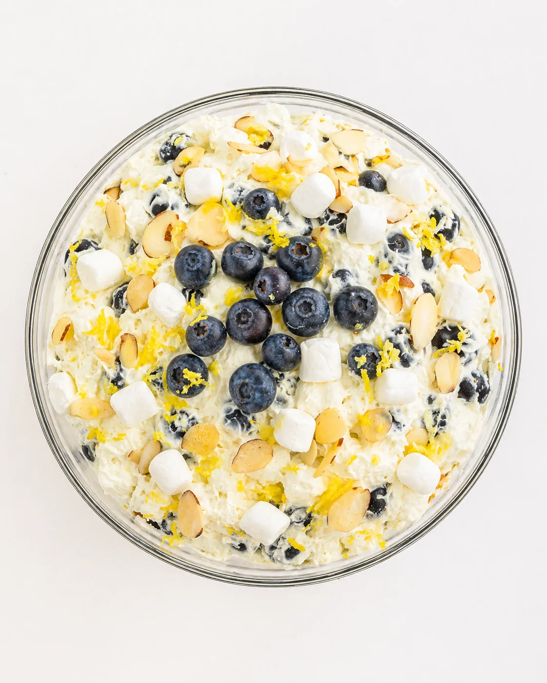 Overhead view of a creamy ambrosia dessert in a glass bowl, topped with blueberries, mini marshmallows, almonds, and lemon zest.