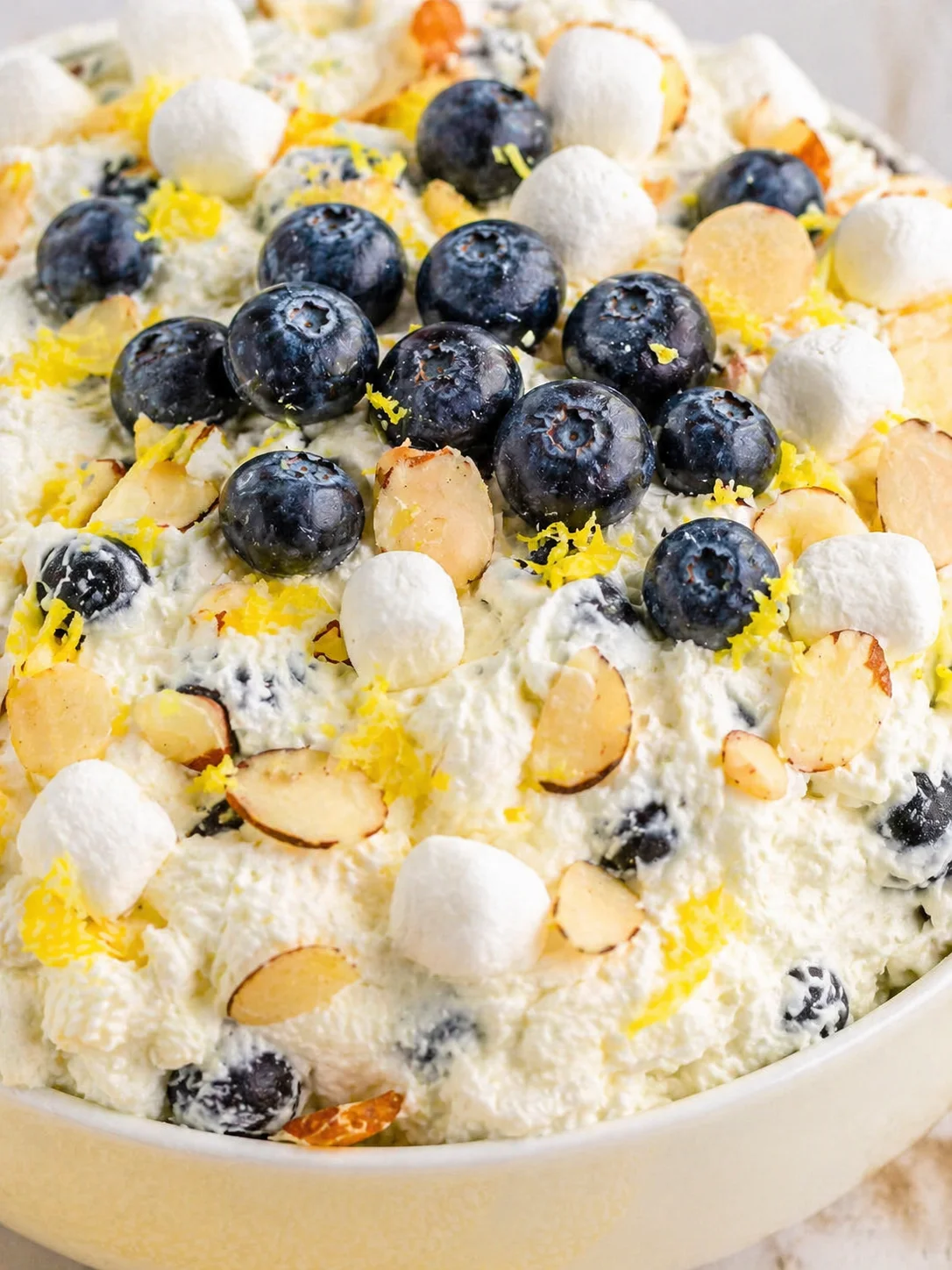 A creamy blueberry-lemon dessert in a pale yellow bowl topped with blueberries, lemon zest, almond slices, and white meringues.