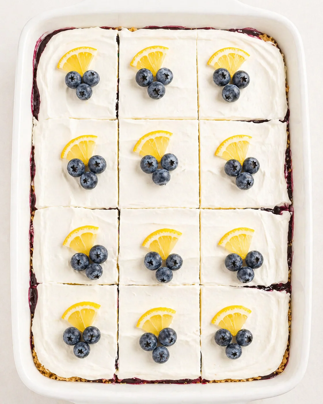 twelve frosted citrus-blueberry dessert squares in a white baking dish, each topped with an orange slice and blueberries.