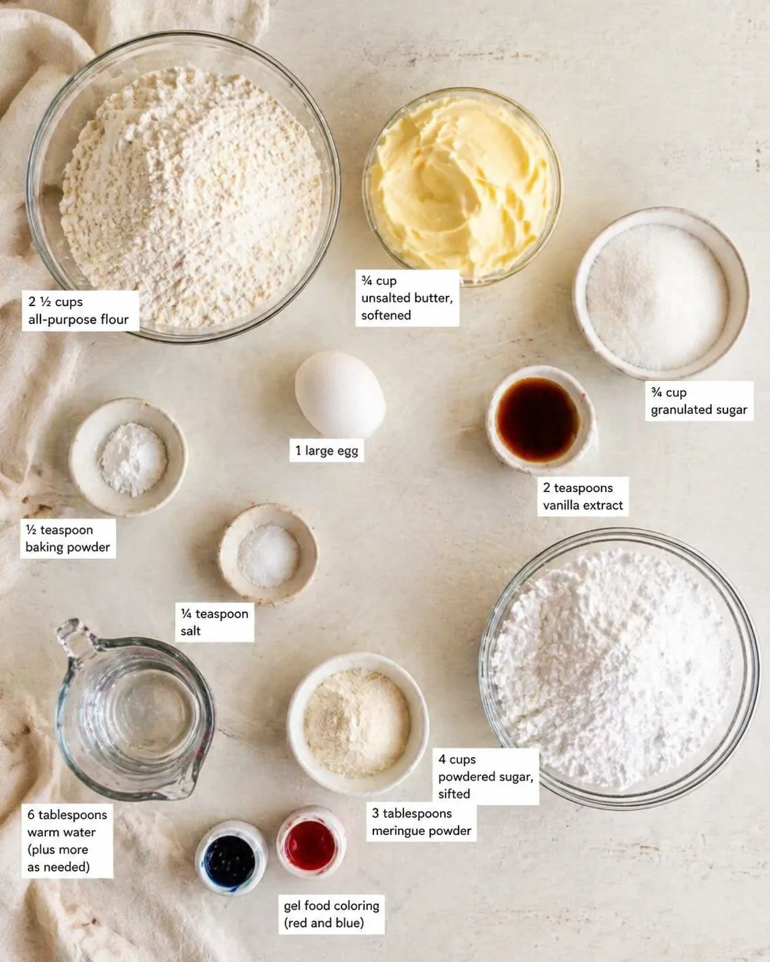 Overhead view of flour, butter, sugar, egg, and powdered sugar with labels on a pale surface.