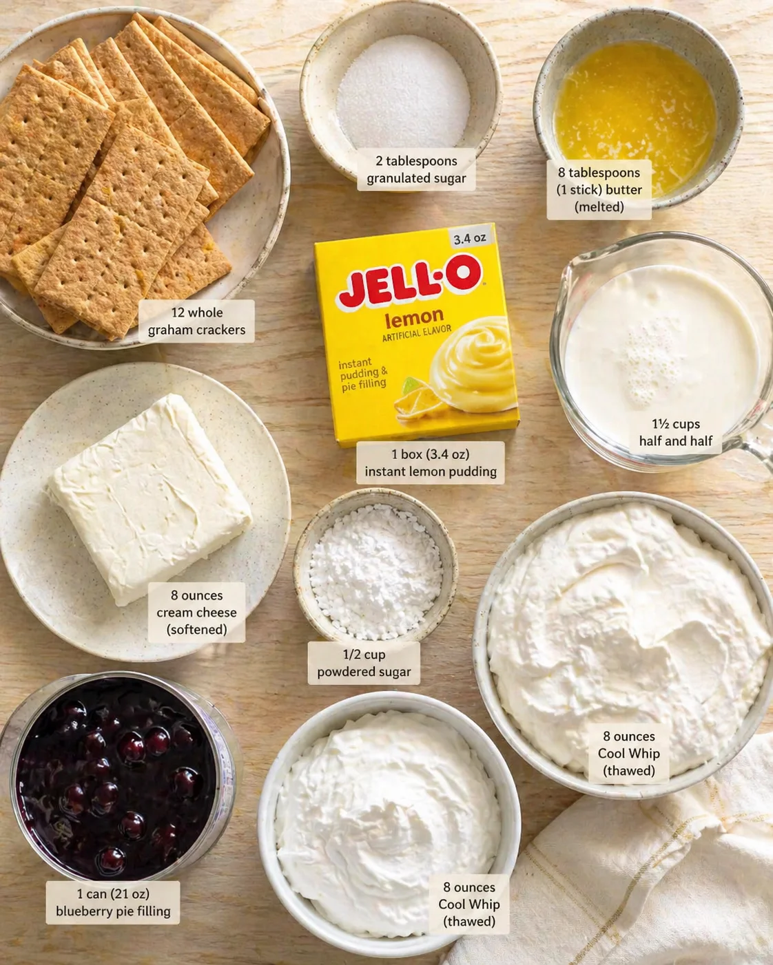 Overhead view of lemon-pie ingredients arranged on a wooden table, including graham crackers, cream cheese, pudding mix, sugar, butter, half-and-half, blueberry pie filling, and whipped topping.