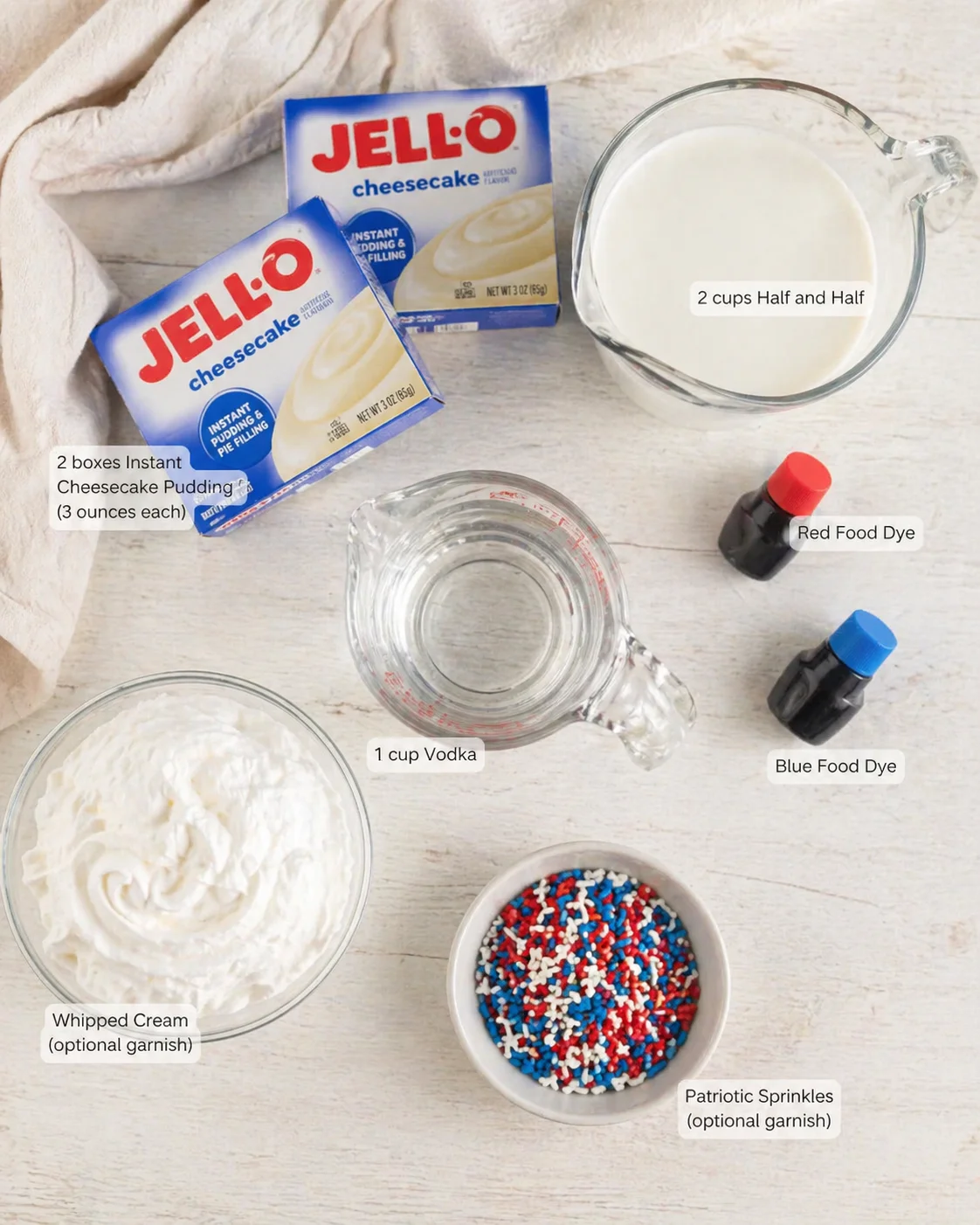 Overhead shot of cheesecake ingredients on a light wooden surface, including Jell‑O boxes, whipped cream, sprinkles, dye bottles, and vodka.
