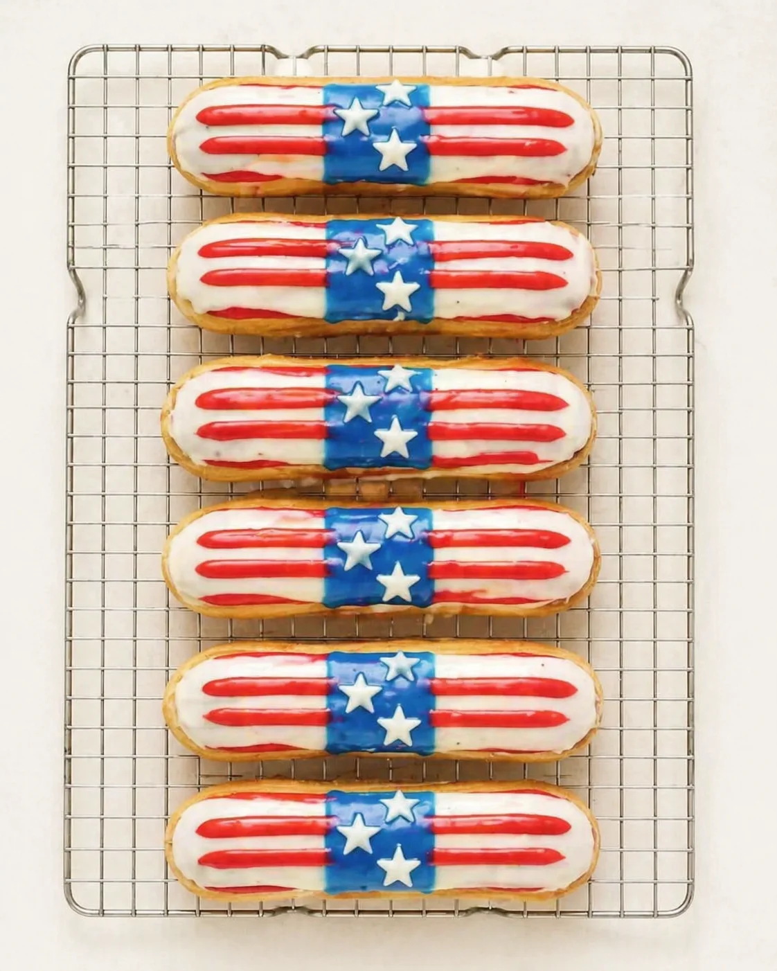 Elaborate patriotic éclairs with red stripes, white stars on a blue band, arranged on a metal cooling rack.