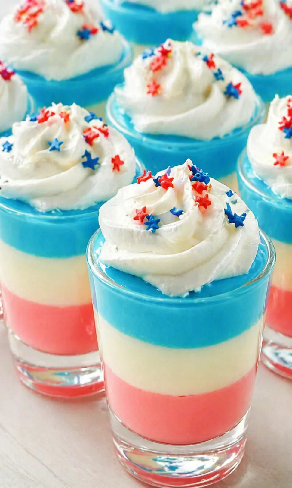 colorful layered dessert shots in clear glasses topped with whipped cream and red-blue star sprinkles.