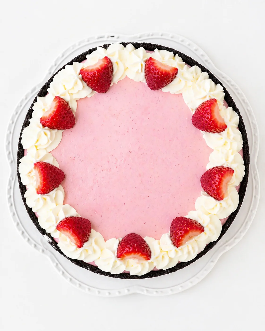 Round cake with pink frosting, white cream rosettes around the edge, and halved strawberries on top.
