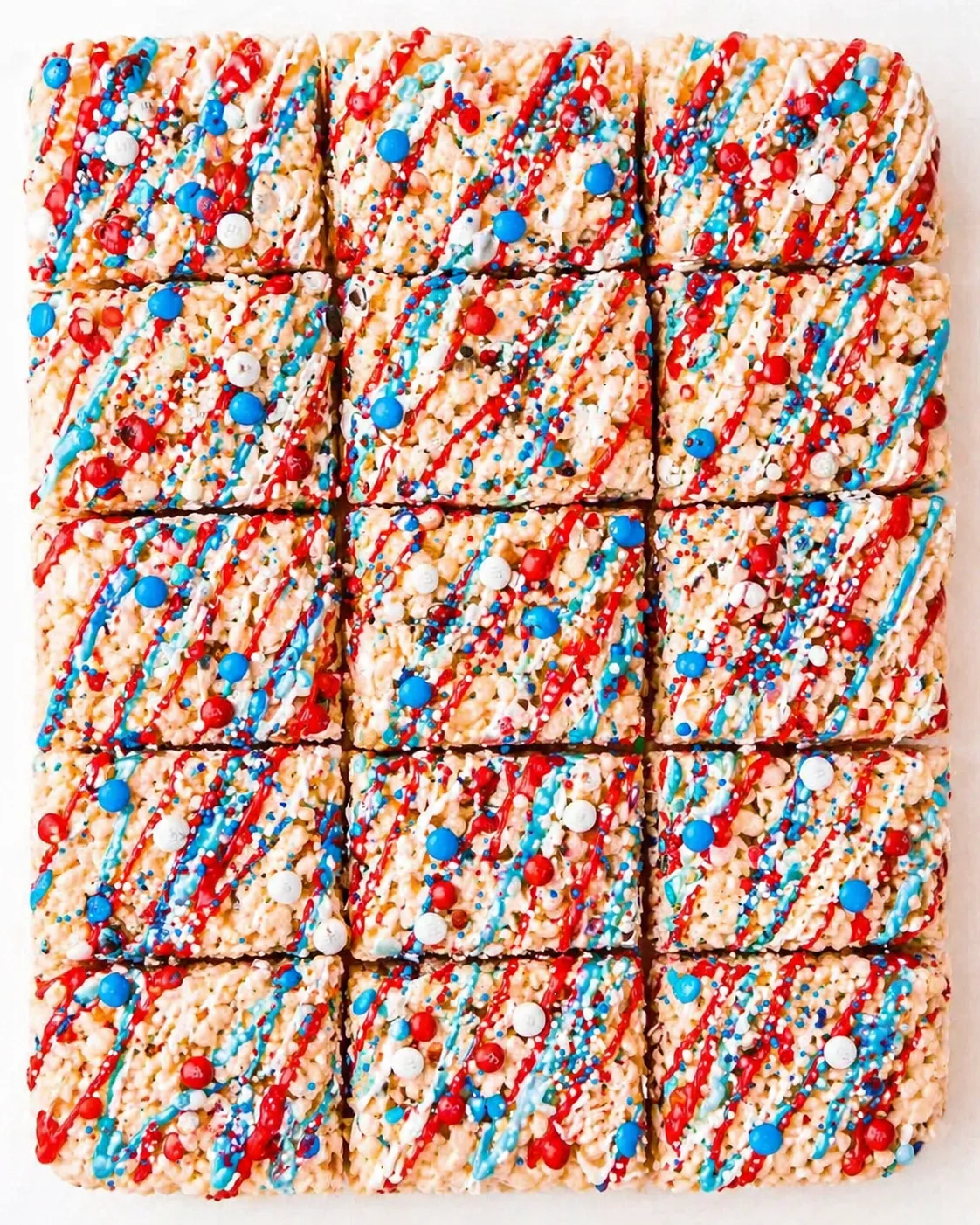 Top-down view of sixteen square Rice Krispie treats on a white tray, decorated with red and blue drizzle and sprinkles.
