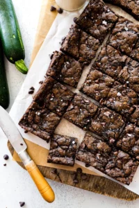 zucchini brownies cut into squares on wood board with knife.