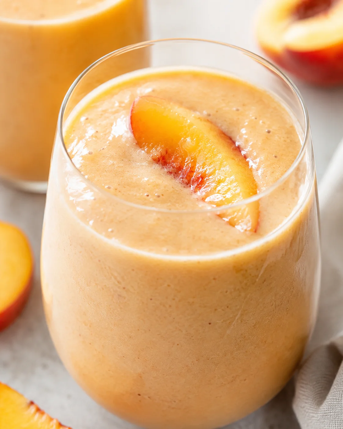 A creamy peach smoothie topped with a fresh peach slice in a clear glass