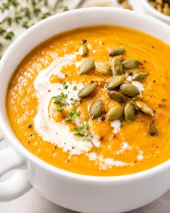 Bright orange carrot soup in a white bowl, swirled with cream and topped with pumpkin seeds and thyme.