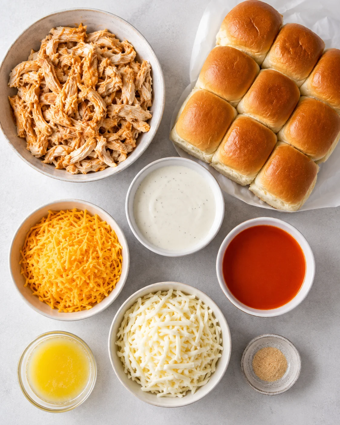 Top-down view of ingredients for buffalo chicken sliders including shredded chicken, Hawaiian rolls, shredded cheeses, ranch, buffalo sauce, melted butter and a