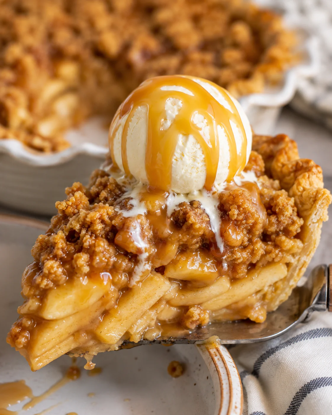 Slice of caramel apple pie topped with vanilla ice cream and caramel sauce on a pie server