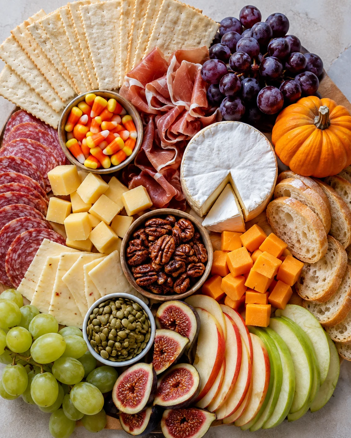 Top-down layout of ingredients for a Thanksgiving charcuterie board including crackers, salami, cheeses, figs, grapes, apple slices and candy corn