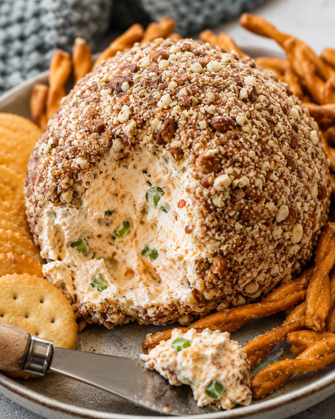 Classic cheese ball coated in chopped pecans with a scoop taken out, served with crackers and pretzel sticks