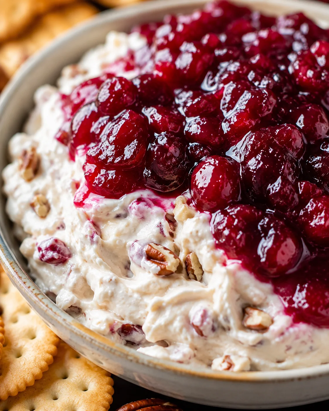 Close-up of cranberry cream cheese dip showing shiny cranberry topping, creamy base and pecans with crackers at the edge