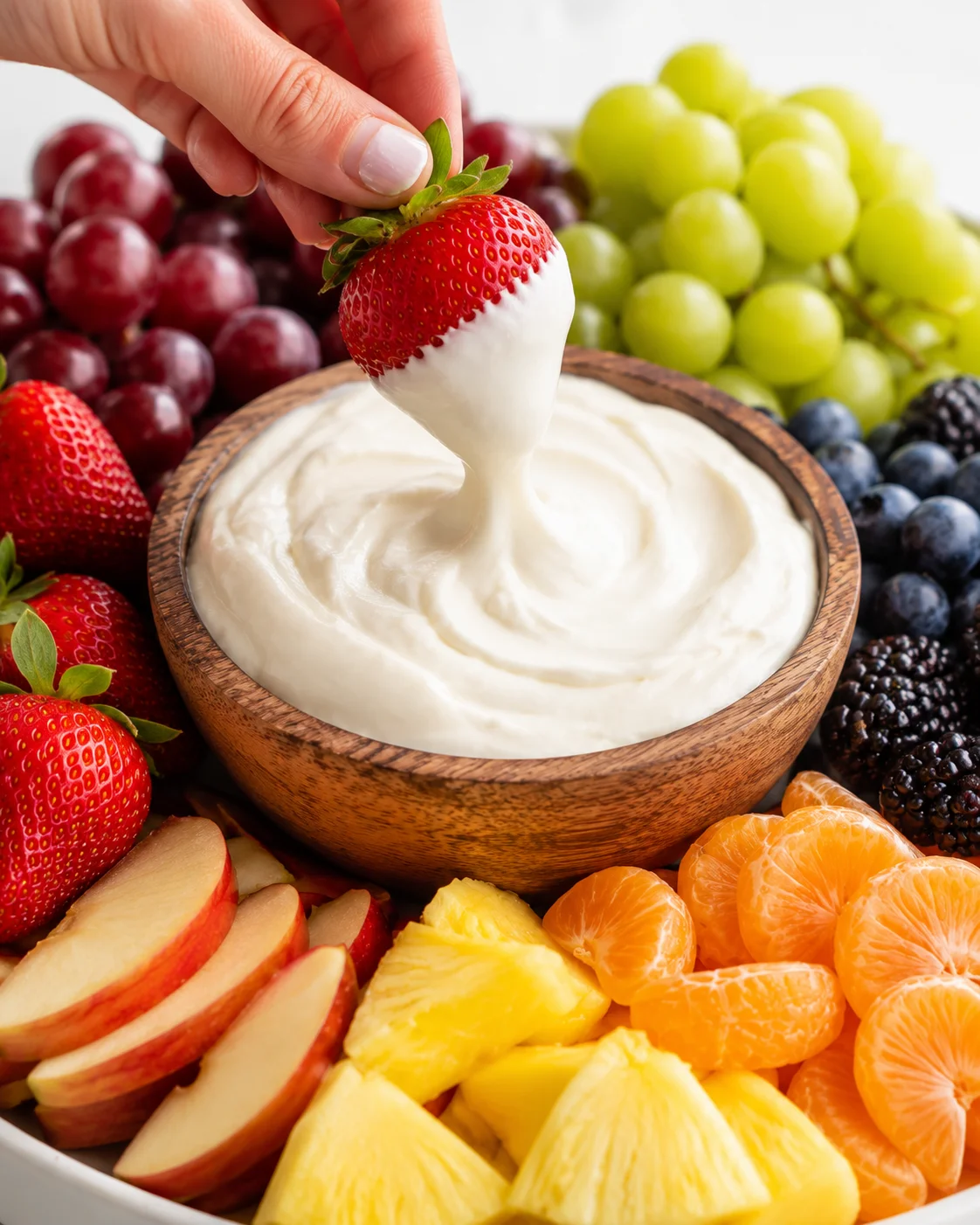 Hand dipping a strawberry into cream cheese fruit dip on a fruit platter