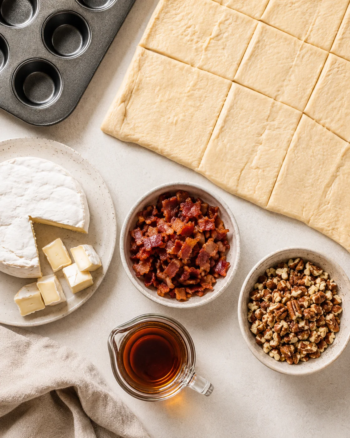 Top-down view of crescent dough, a wheel of brie, a bowl of cooked diced bacon, chopped pecans and a small pitcher of maple syrup