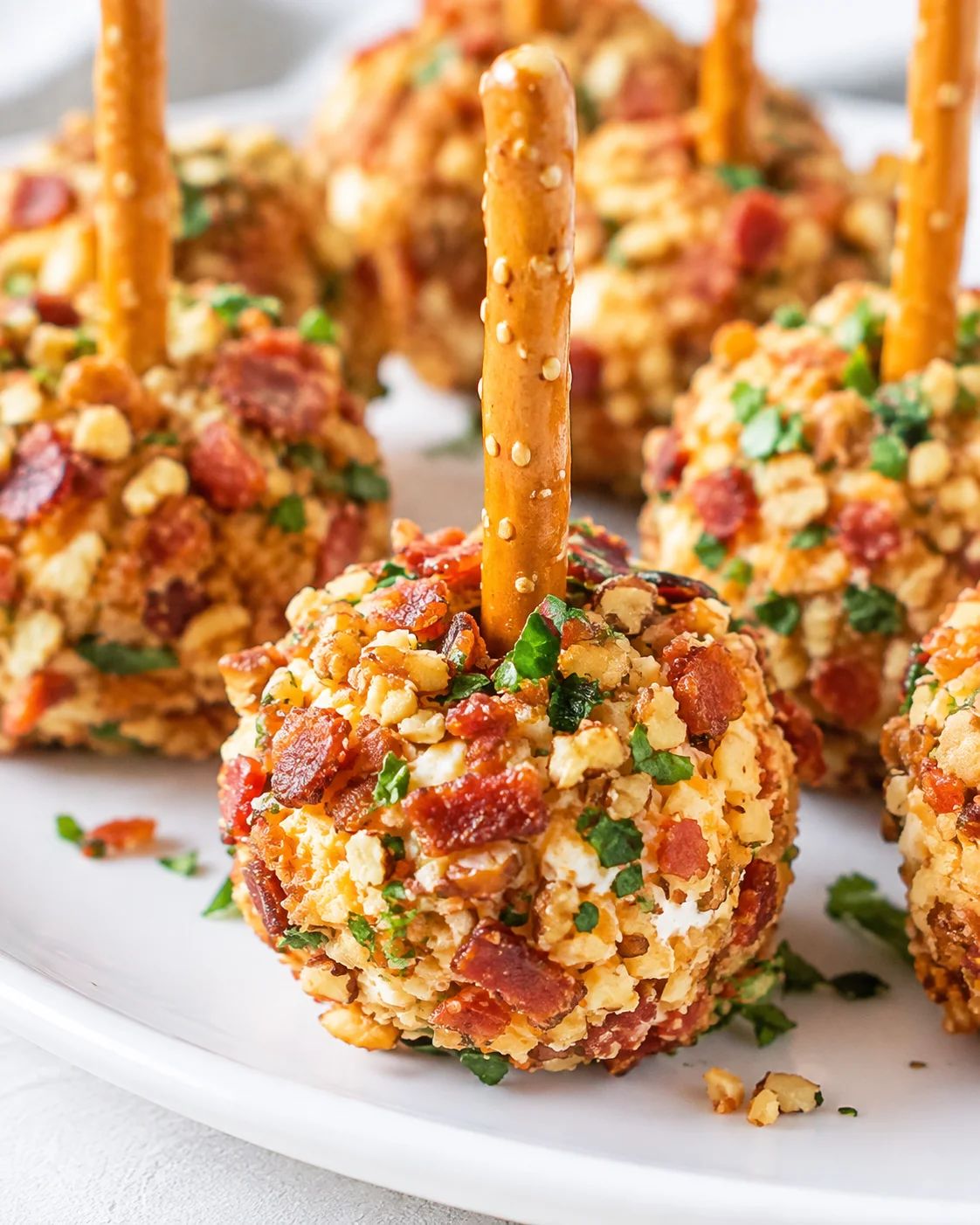Close-up of a mini cheese ball coated in bacon and chopped nuts with a pretzel stick, on a white plate