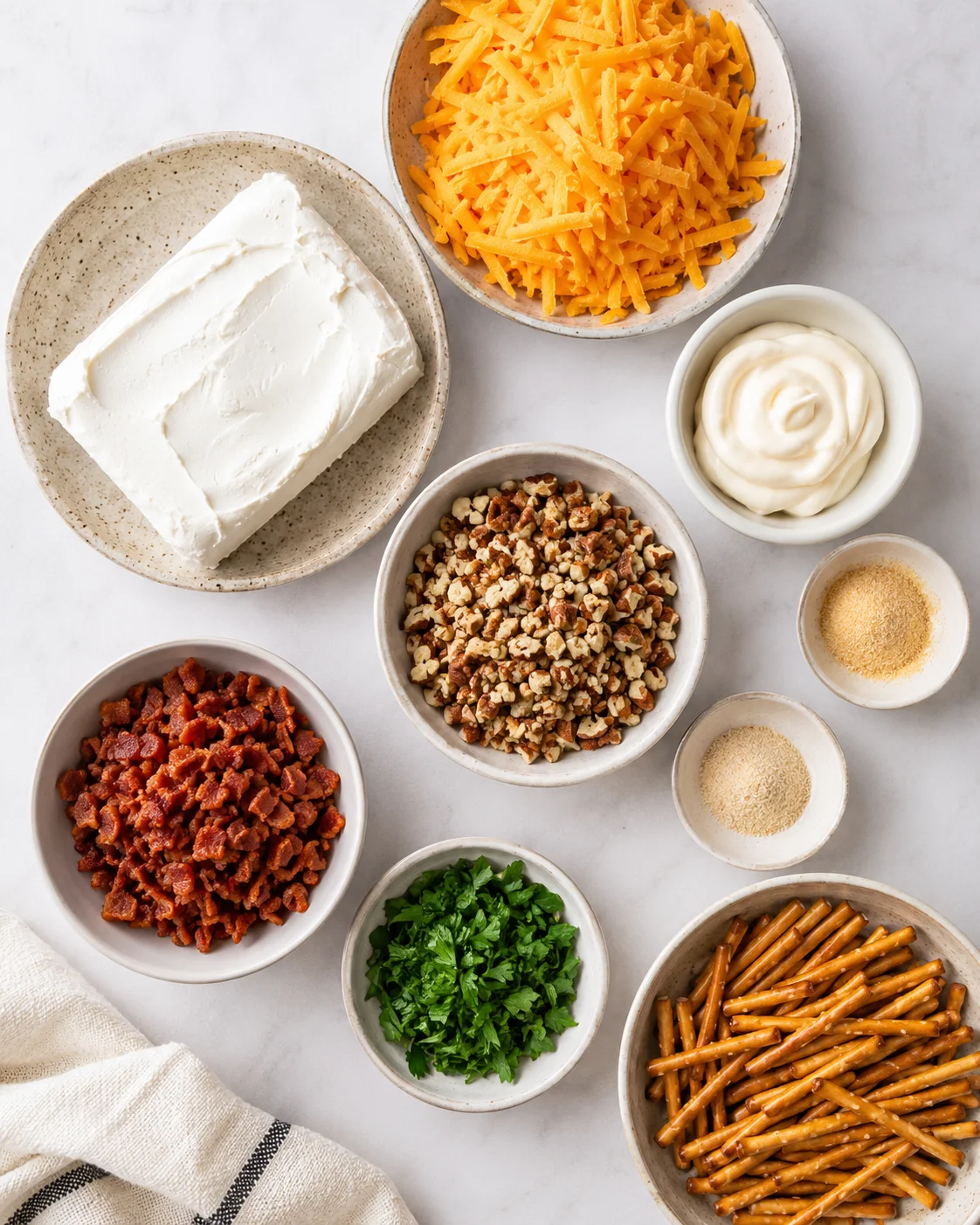Top-down view of bowls with cream cheese, shredded cheddar, mayonnaise, chopped nuts, bacon bits, parsley, pretzel sticks and seasonings