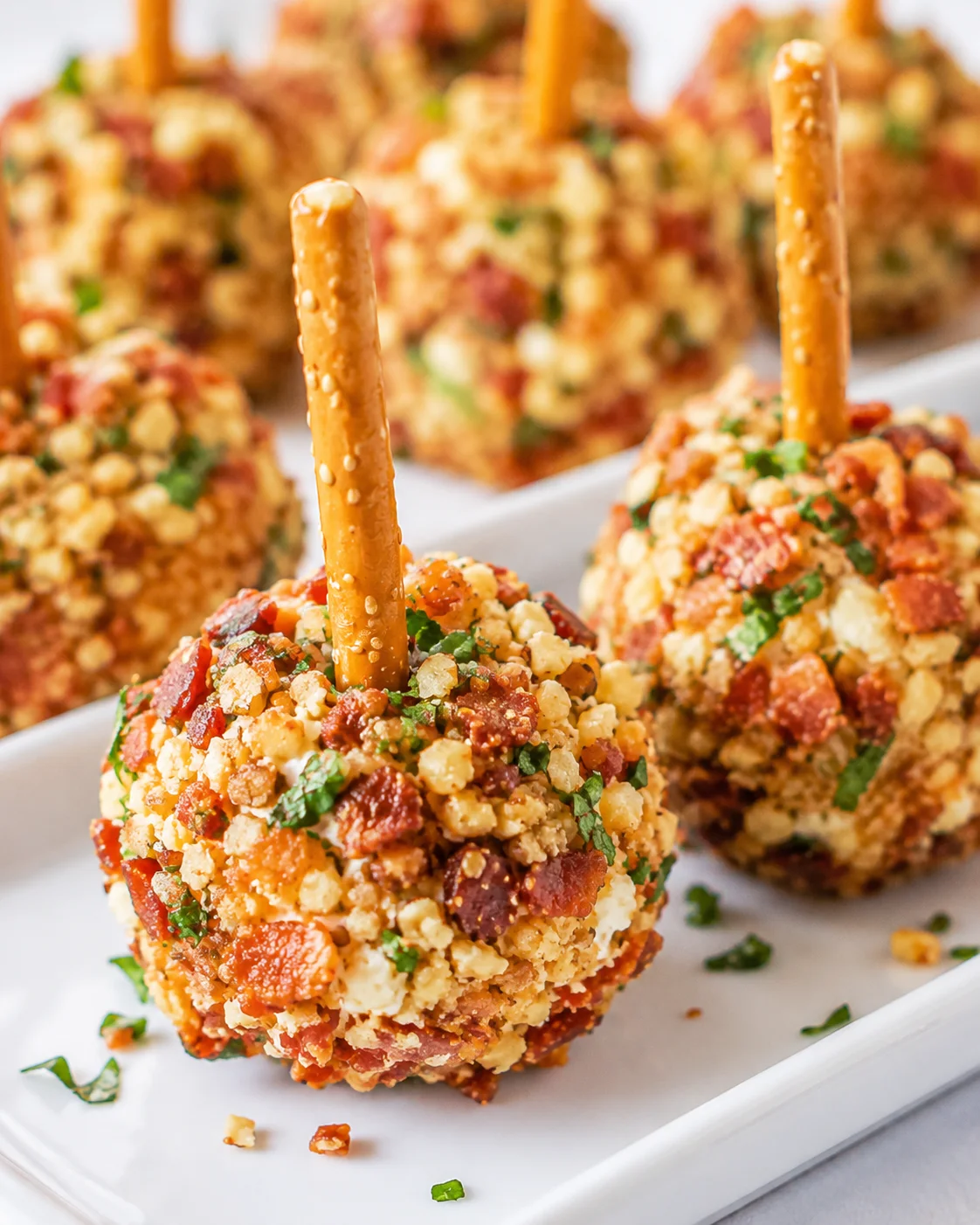 Side view of mini cheese balls coated in bacon and nuts arranged on a white platter with pretzel sticks