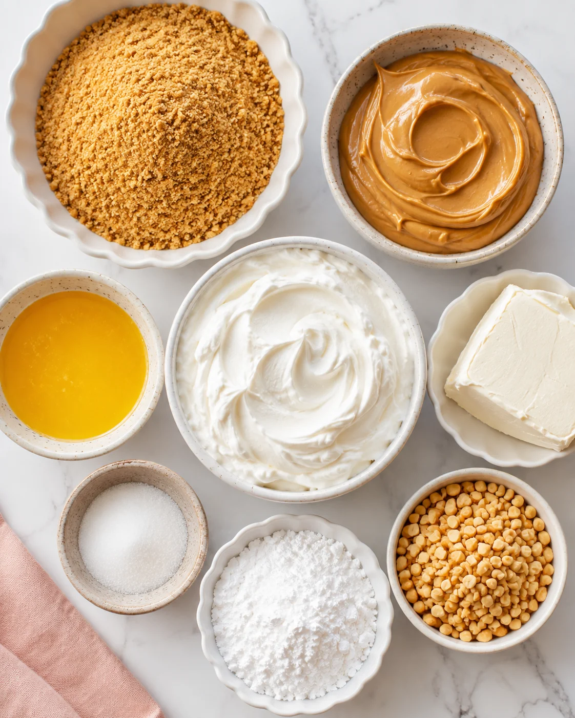 Top-down view of bowls with graham crumbs, peanut butter, cream cheese, whipped cream, melted butter, powdered sugar and chopped peanuts