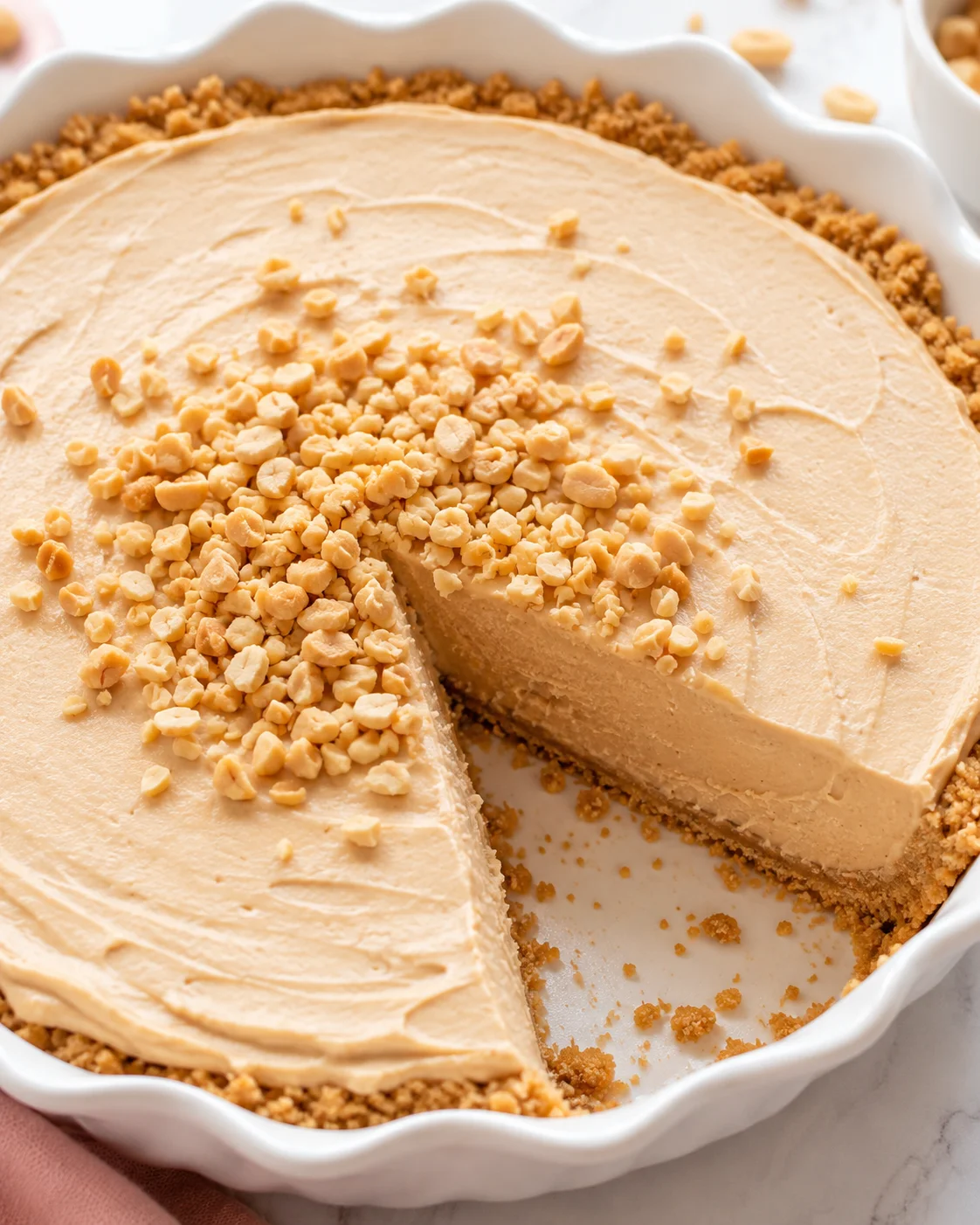 Whole no-bake peanut butter pie in a white fluted dish with chopped peanuts on top and one slice removed