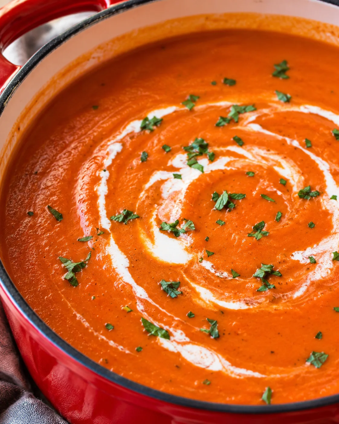 A rich and smooth tomato bisque garnished with fresh herbs in a red pot.