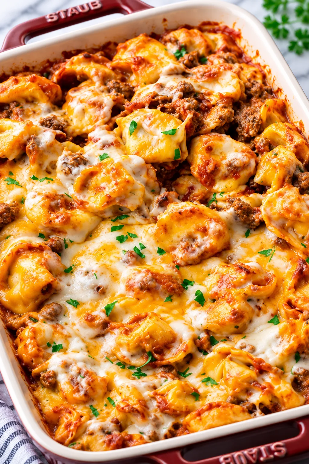 Cheesy baked tortellini with tomato meat sauce in a red Staub dish, sprinkled with parsley.