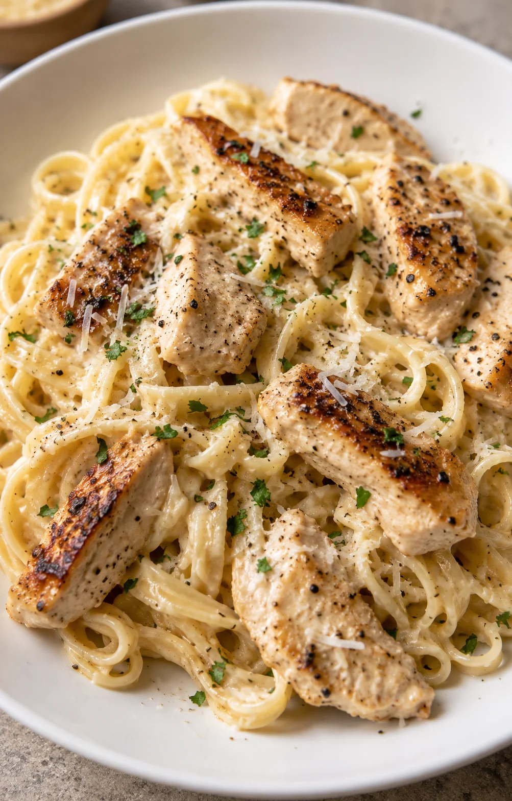 Creamy fettuccine alfredo topped with grilled chicken in a white bowl, garnished with parsley.
