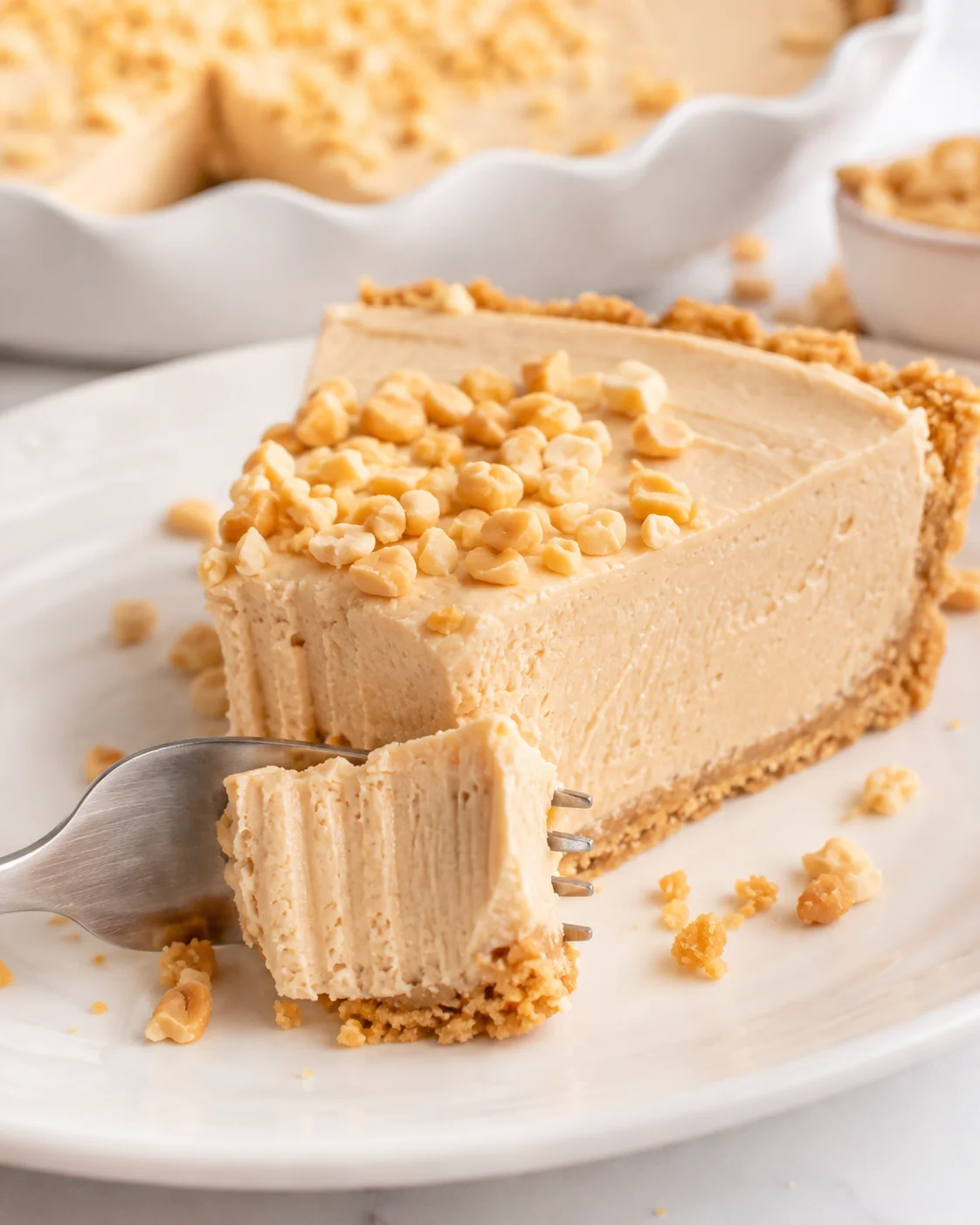 Slice of no-bake peanut butter pie on a white plate with a fork lifting a bite, pie dish blurred in the background