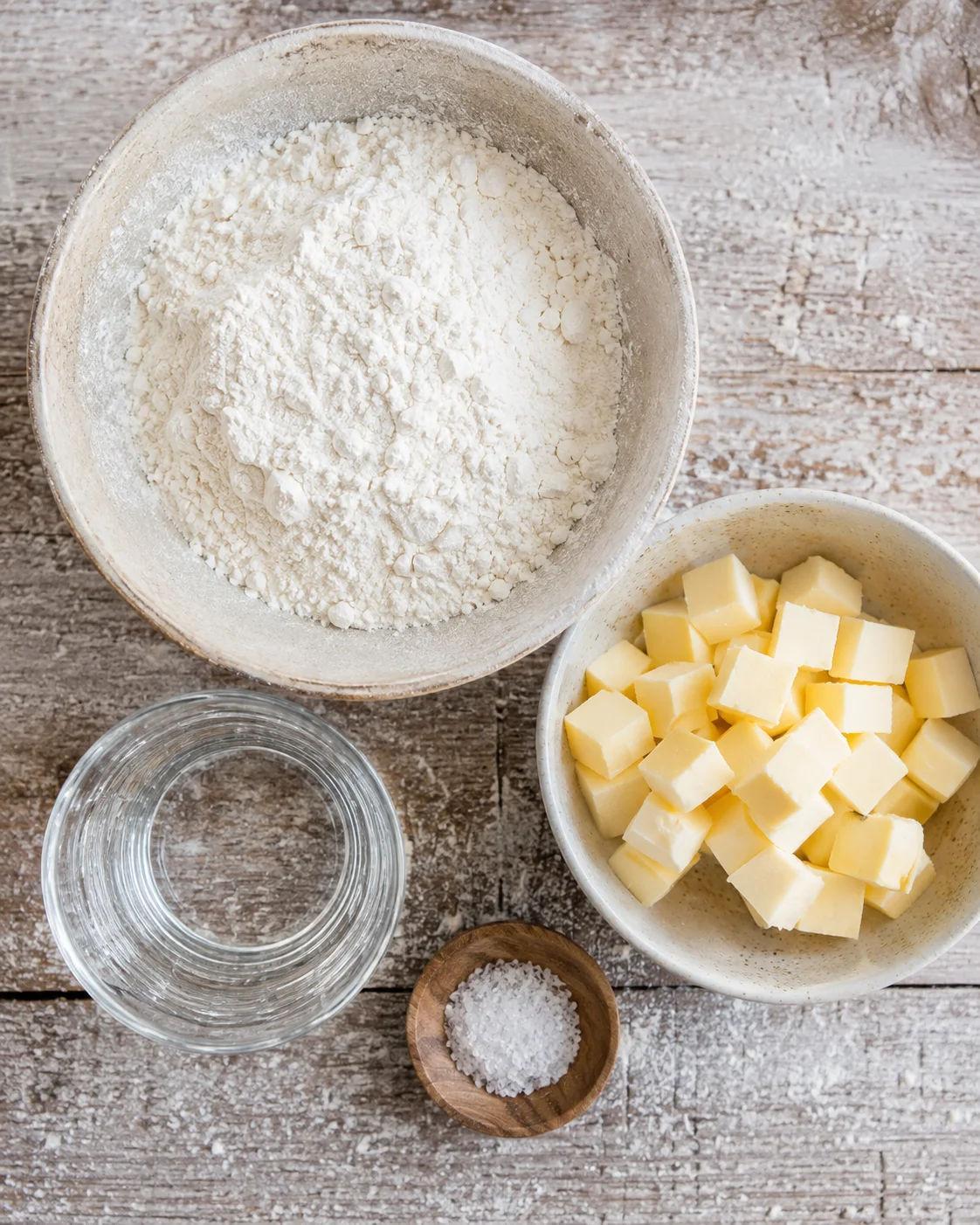 Top-down view of flour, cubed butter, a glass of water and salt arranged on a wooden surface for puff pastry.