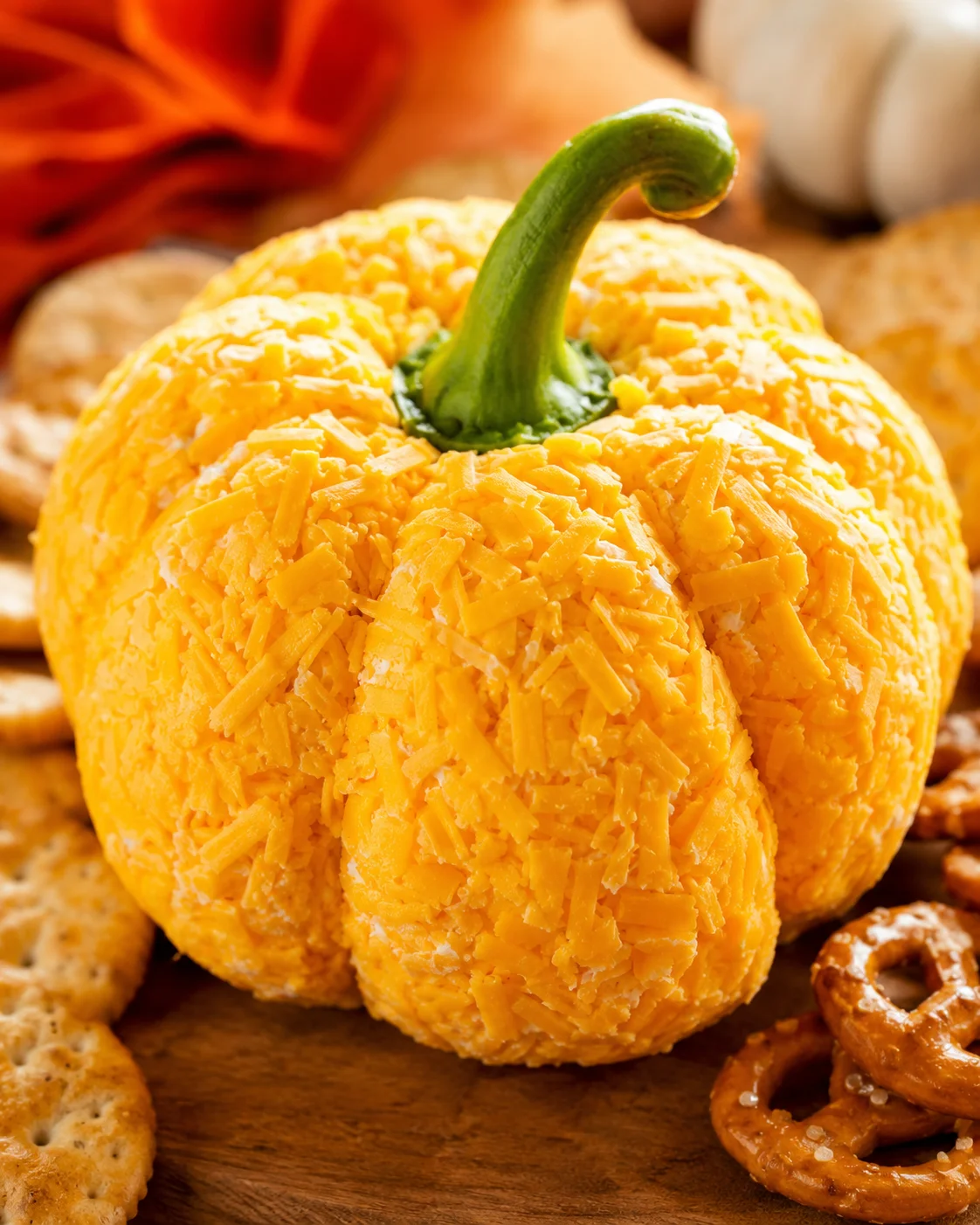 Pumpkin-shaped cheese ball coated in shredded cheddar on a wooden board with crackers and pretzels