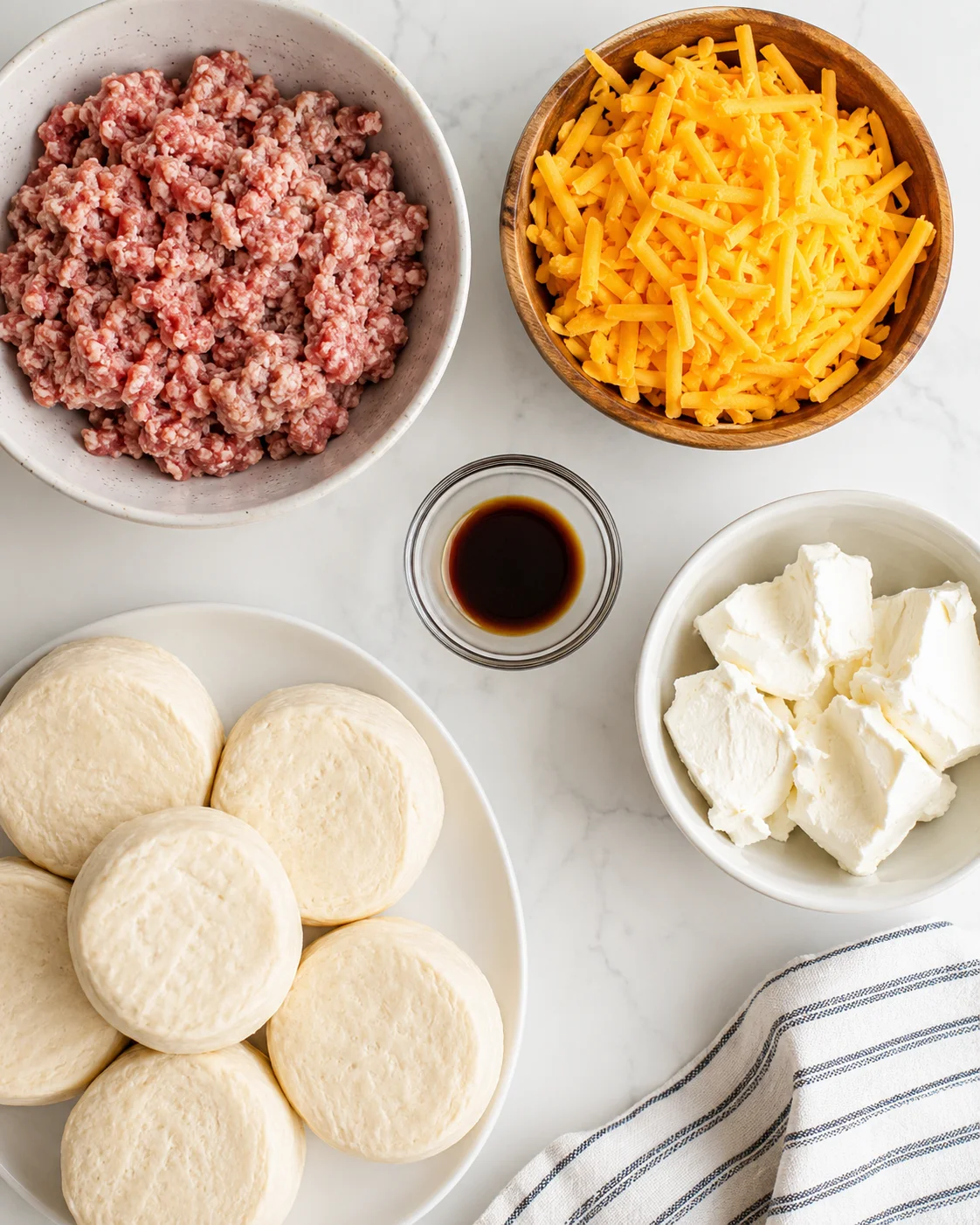Top-down view of raw sausage, shredded cheddar, cream cheese, biscuits, and Worcestershire sauce
