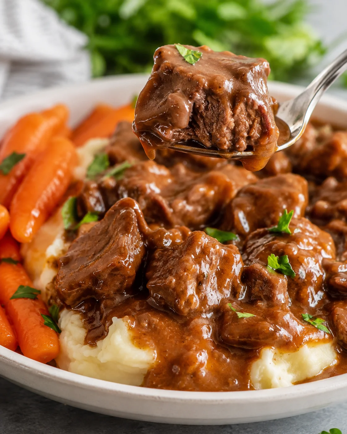 Fork holding a tender cube of slow cooker beef tips with gravy over mashed potatoes and carrots