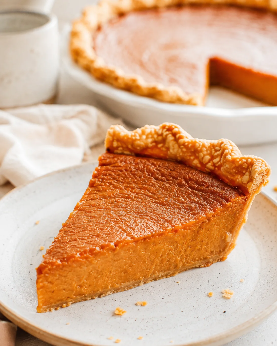 Side view of a single slice of sweet potato pie on a ceramic plate with the remaining pie blurred in the background