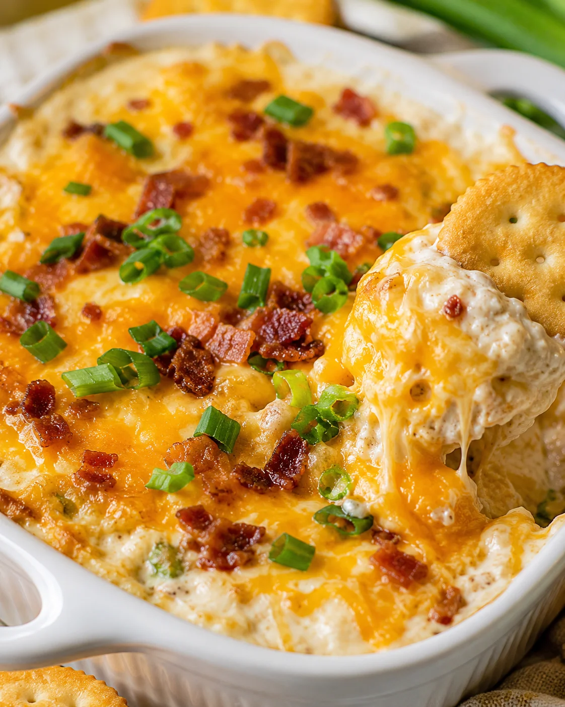 Warm & Cheesy Crack Dip in a white baking dish with melted cheddar, bacon bits and green onions and a cracker dipping into the cheese