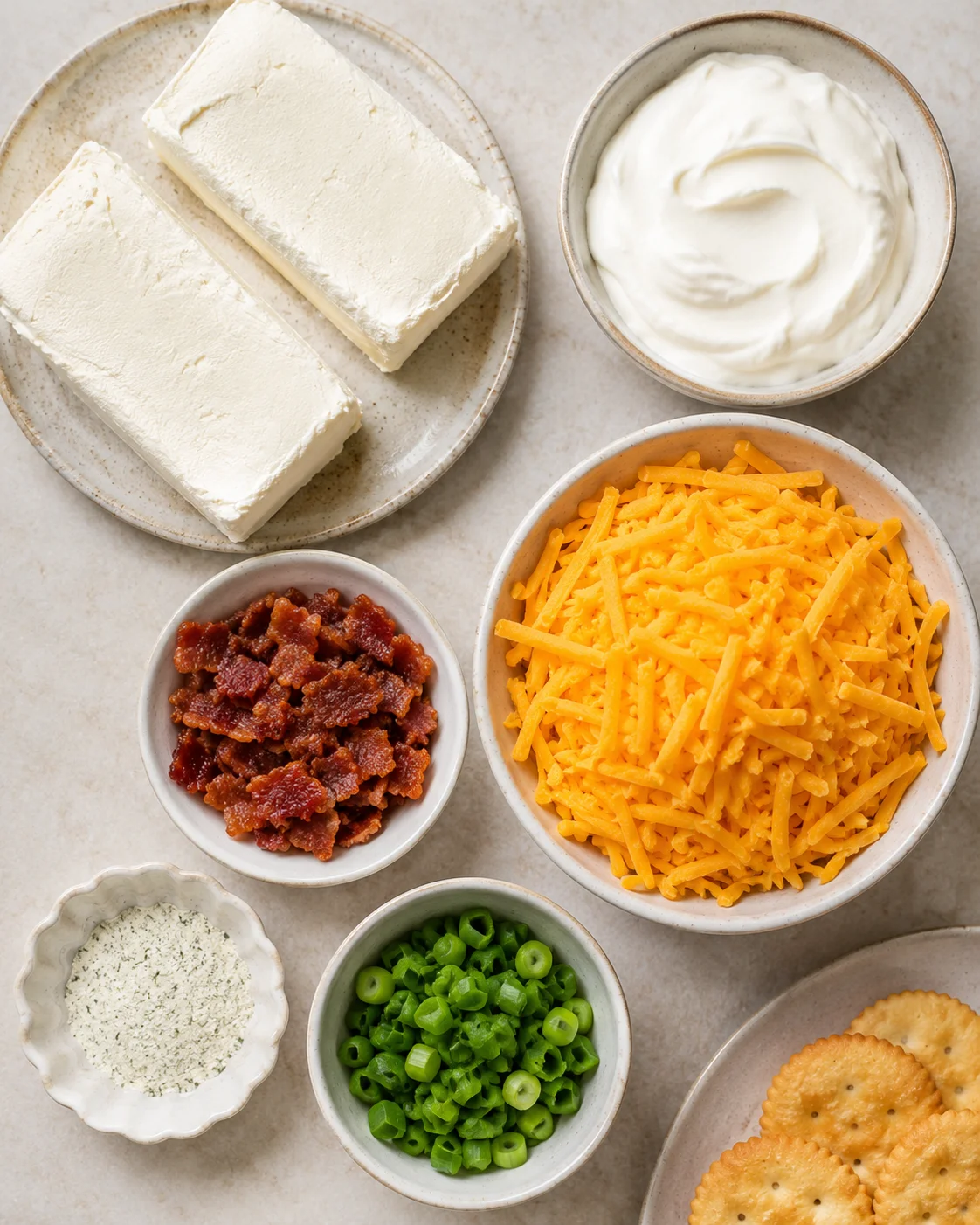 Top-down view of ingredients for Warm & Cheesy Crack Dip including cream cheese blocks, sour cream, shredded cheddar, bacon bits, ranch mix and sliced green on-